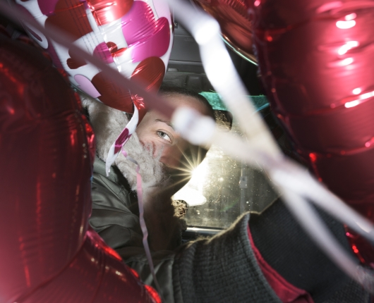 A man in the driver's seat turning around to look at the camera lens through a cluster of balloons.