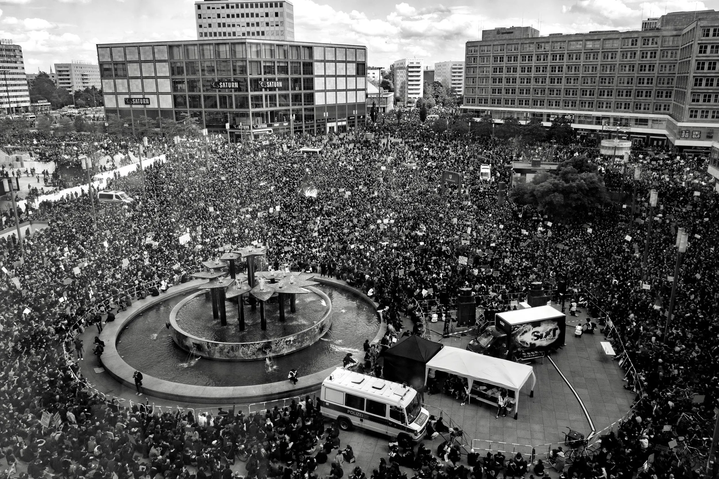A massive crowd filling a city center with a fountain. 