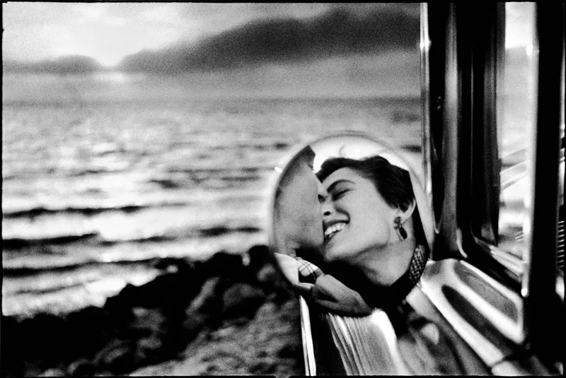 A woman about to kiss a man seen in the car's side mirror. 