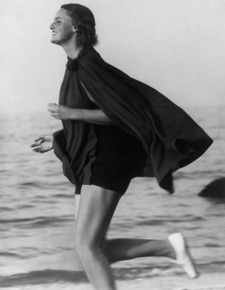 A woman in a one piece swimsuit with a cape running on the beach.