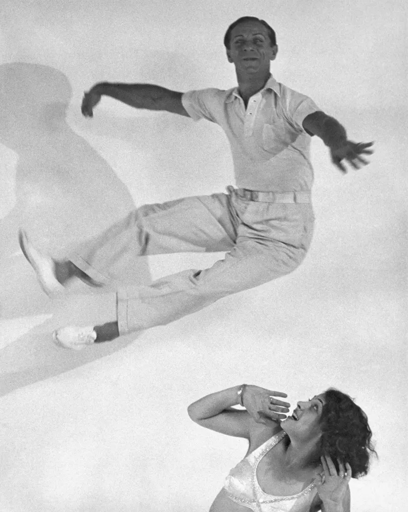 A man jumping through the air, as a woman beneath him looks up in performative alarm. 