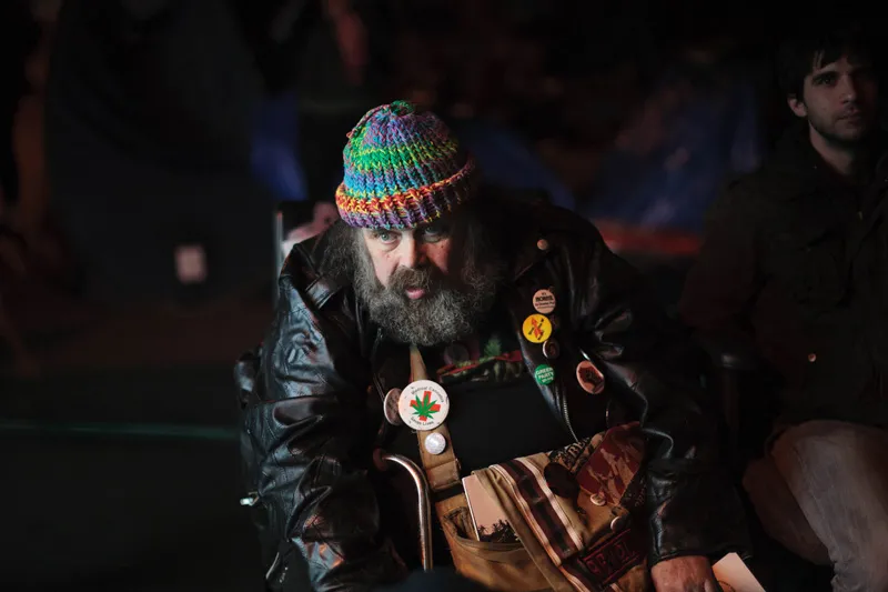 A disgruntled older man in a beard with a leather jacket looking on. 