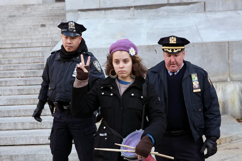 A young woman raising the peace symbol as she is being escorted by two cops.