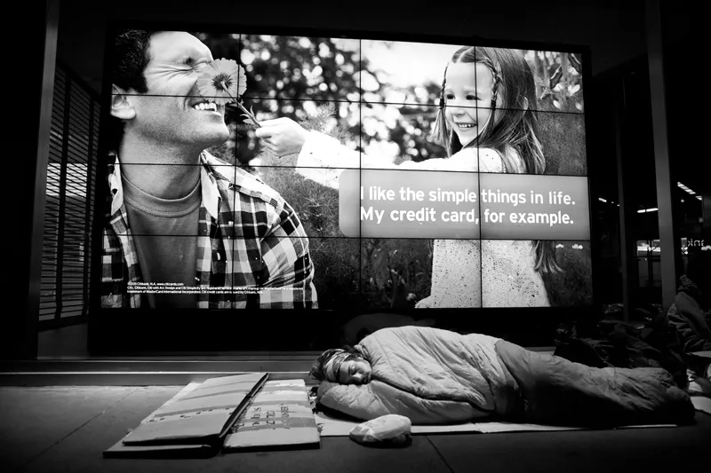 A homeless man sleeping on the streets bathed in the glow of a credit card advertisement. 