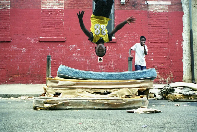 A young black boy backflipping onto a blown out mattress in the middle of the street. 