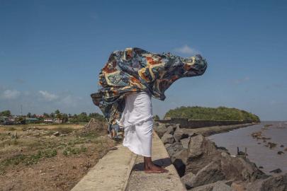 Black person in flowing clothing blowing in the wind holds arms out, face obscured.