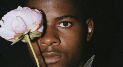 A man in the dark, with a flower raised and covering one eye. 