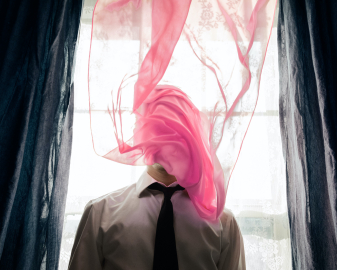 A man standing in front of the bright window, an almost transparent pink curtain wrapped around his face.