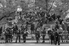 A large crowd sitting on rocks and leaning against a police barricade watching something out of frame.