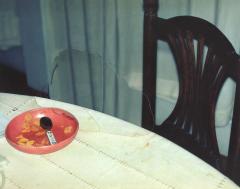 A dining room chair and table on which an abandoned a bowl of partially eaten cereal remains.