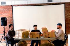 Three people in front of a screen with boxes on the table. 
