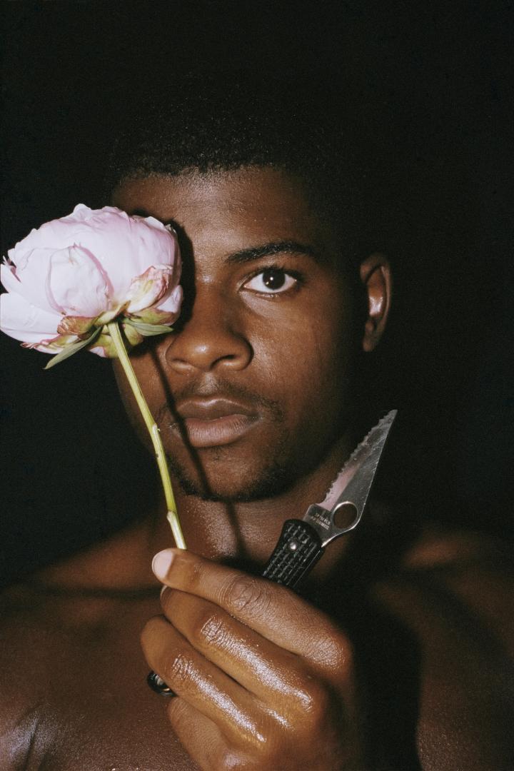 A shirtless man with a flower and a knife in his hand. 
