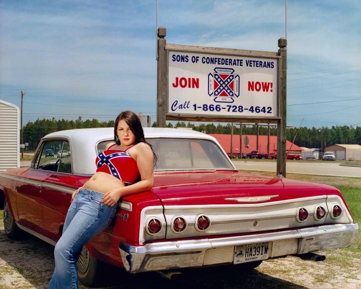 A woman leaning on a car with a confederate shirt.