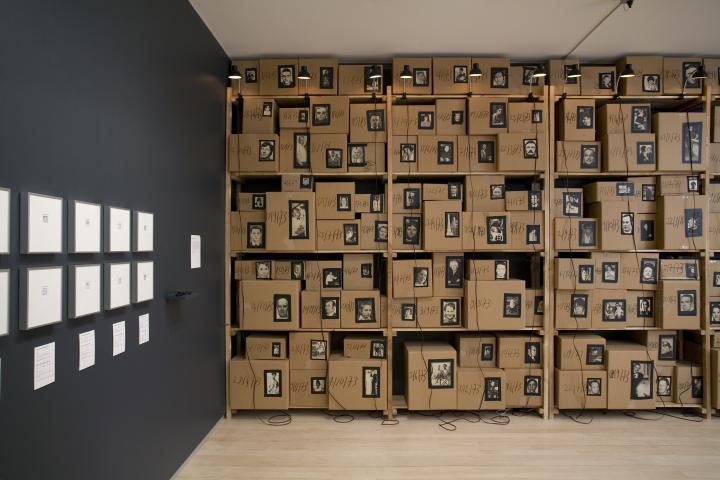 A photography exhibition featuring a wall of cardboard boxes, each one with a respective profile of someone. 