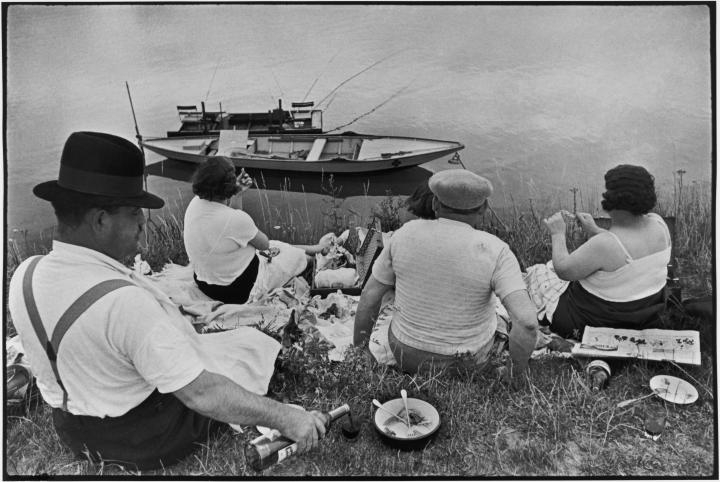 Men and women having a picnic by the side of waterway where a boat is floating. 
