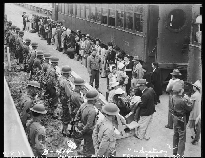 A row of US military soldiers look on while Japanese Americans are shuffled to internment camps. 