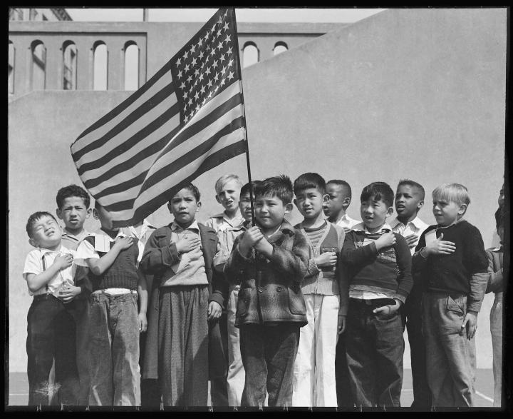 Two rows of young boys reciting the national anthem with their hands on their hearts, one waving the American flag above his head. 