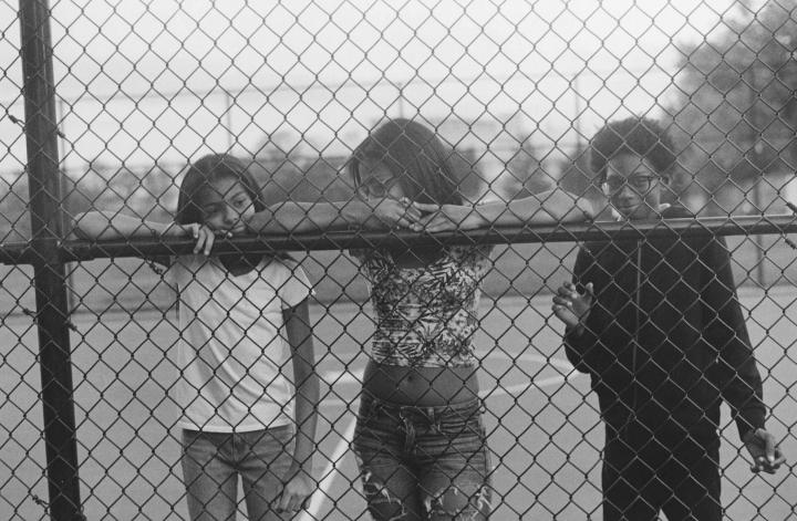 Three youths standing on a basketball court behind a chain link fence. 