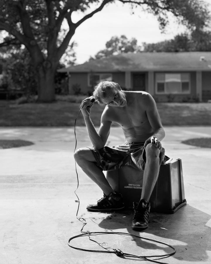 A shirtless man sitting on a recycling bin in the middle of a suburban street shaving his head. 