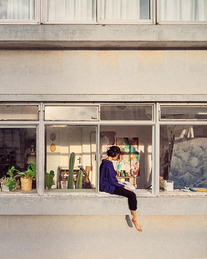 A woman on the windowsill of her apartment reading, her foot dangling in the open air. 