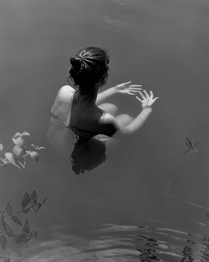 A woman crouched in water, looking at her hands submerged in the water. 