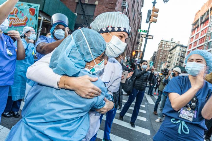 Healthcare workers in the middle of a New York City street embrace, as people applaud them. 