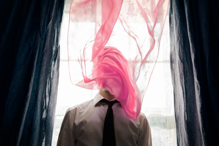 A man standing in front of the bright window, an almost transparent pink curtain wrapped around his face.