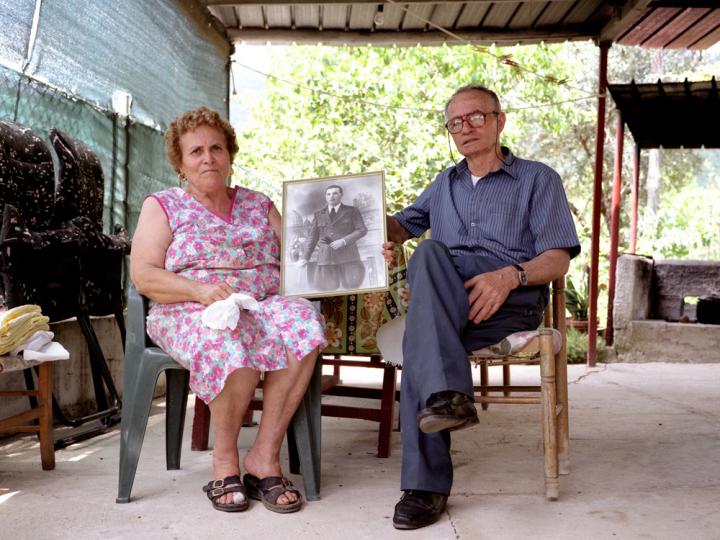 A old couple holding a photo of a young man in a suit and tie. 