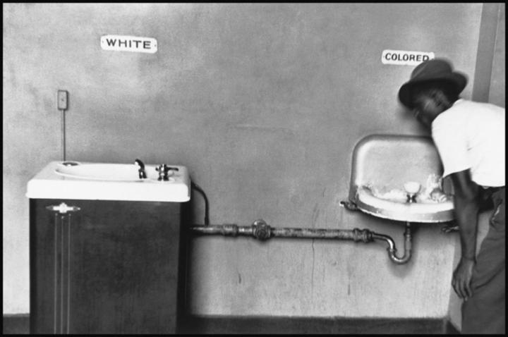 A young black boy drinking from a water fountain for black people only, alongside a whites only water fountain that is more maintained. 