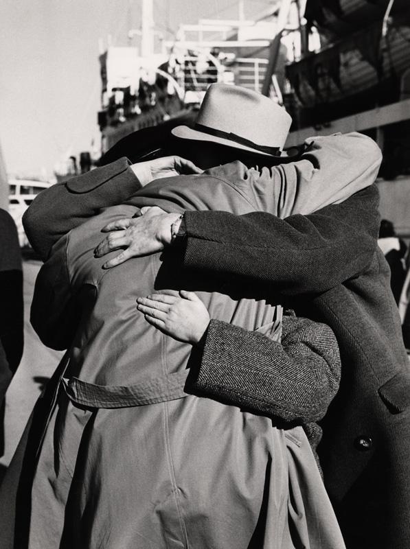 Two people embracing on the docks with a large ship in the background. 