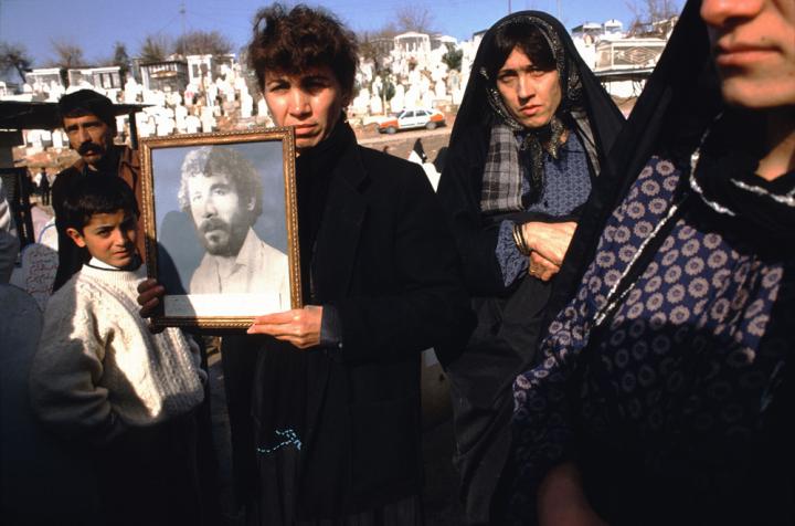 A family in mourning in a graveyard, the wife holding up a framed photo of the departed husband.