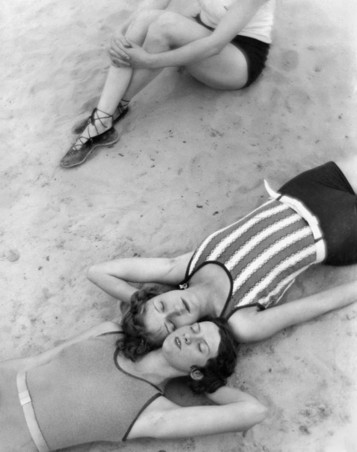 Two women laying on the beach, faces pressed cheek to cheek. 