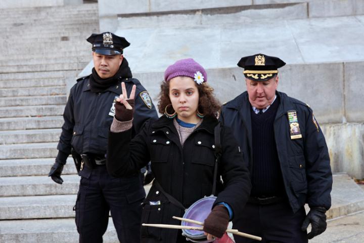 A young woman raising the peace symbol as she is being escorted by two cops.