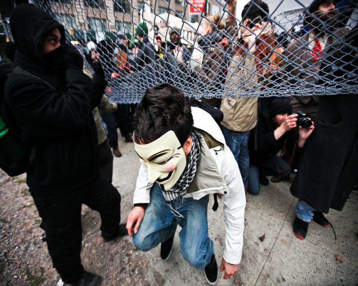 A man in a Guy Fawkes mask crawling under a chain link fence during a protest.