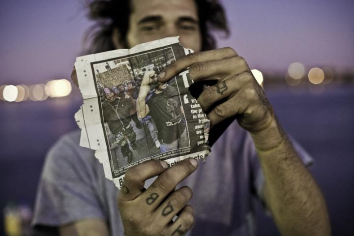 A man with hand tattoos lifting up a torn newspaper, pointing at a picture of protests. 