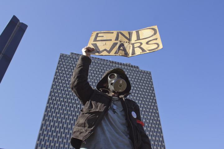 Someone wearing a gas mask standing in front of a skyscraper and they are raising a sign that says, "End Wars". 