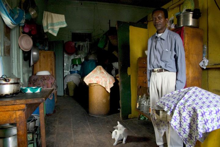 A man standing in his kitchen, while a white cat next to him cleans itself. 