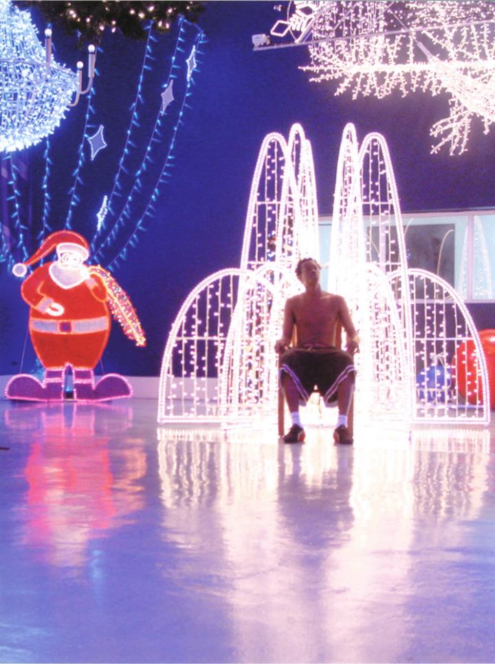 A shirtless man sitting on a chair surrounded by bright Christmas decor. 