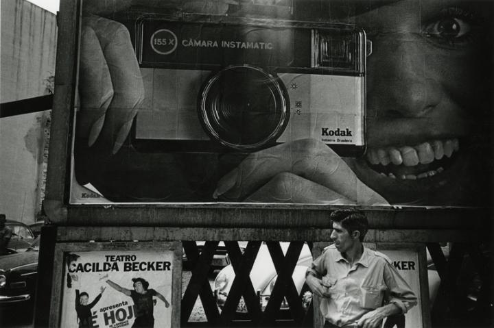 A man digging in his pocket for his lighter with a billboard advertising a Kodak camera behind him.