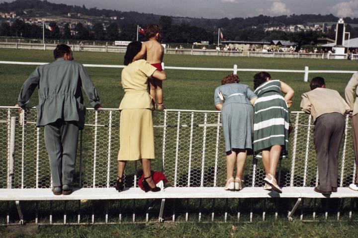A row of people watching a sports event from the sideline, with one woman lifting a child so they can see. 