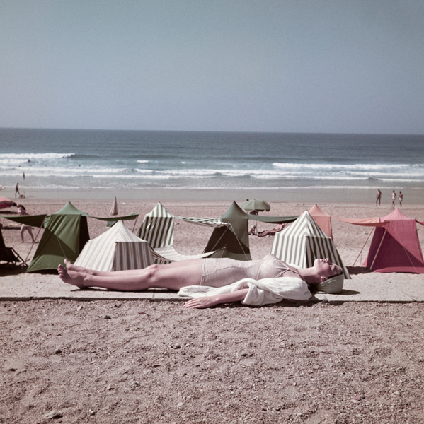 A woman tanning on the beach, with rows of tents behind her. 