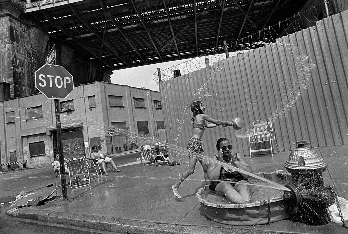 A woman in a kiddie pool next to a open fire hydrant on a inner city street. 