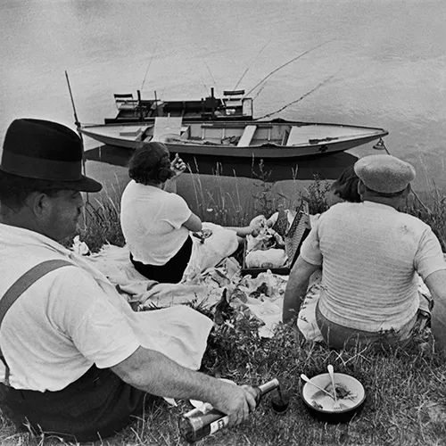 Men and women having a picnic by the side of waterway where a boat is floating. 