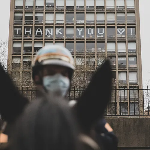 An out of focus police officer with a mask astride a horse, with the camera focusing on the background, an office building where a row of windows spell out "Thank You" alongside a heart with the help of poster boards. 